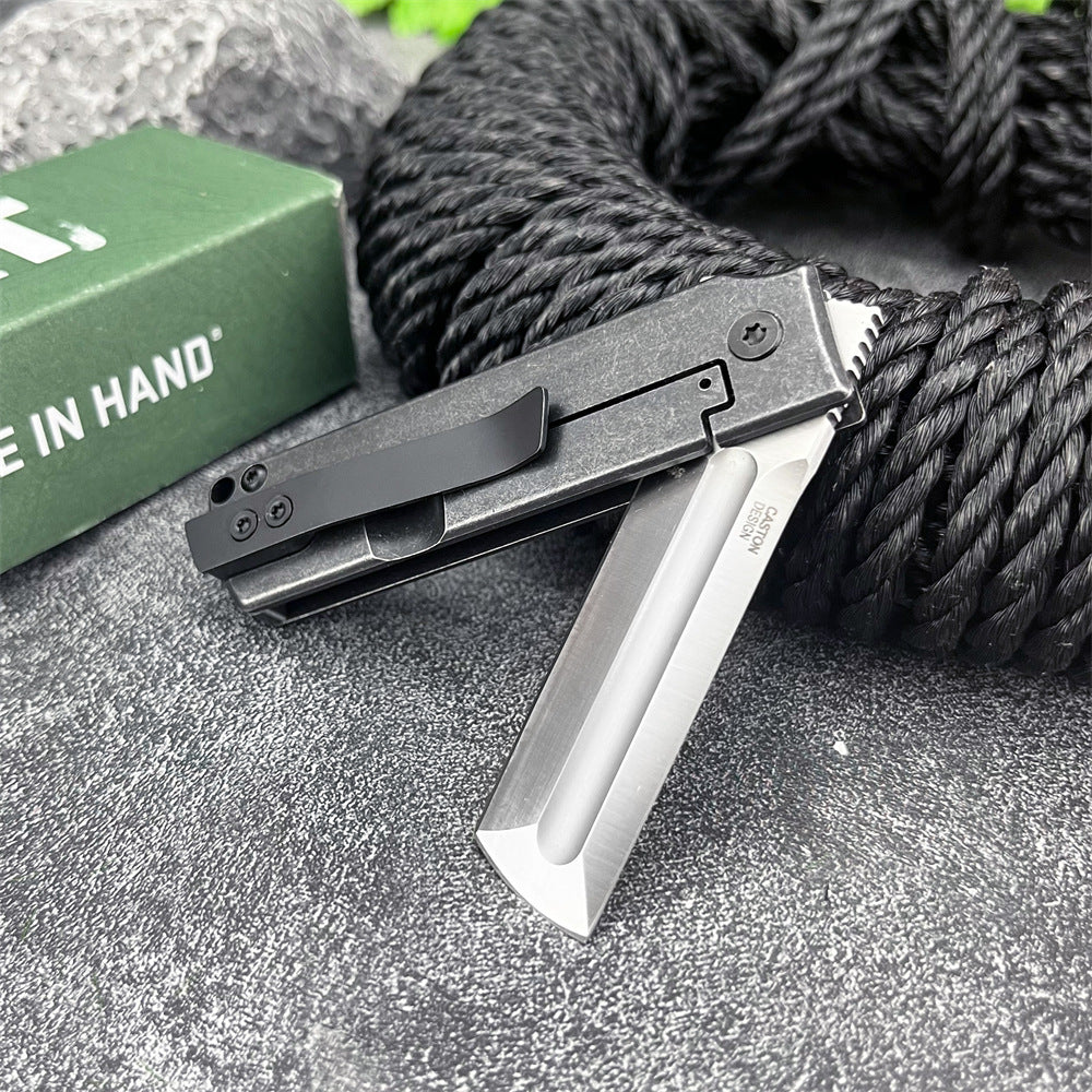 An open CRKT folding knife with a black handle and silver tanto blade, displayed on a grey surface with black rope and a green box.