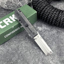 An open CRKT folding knife with a silver tanto blade and black handle, displayed on a grey surface with a green box and black rope.