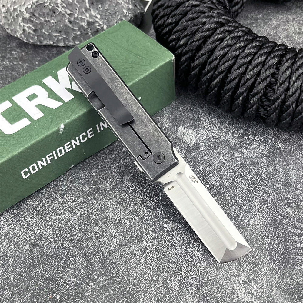 An open CRKT folding knife with a silver tanto blade and black handle, displayed on a grey surface with a green box and black rope.