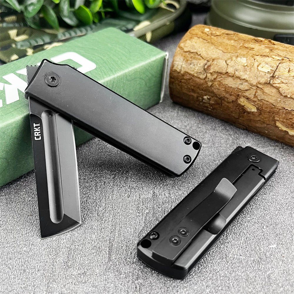 An open and a closed CRKT folding knife displayed on a grey surface with a green box, wood log, green plants, and a camping lantern.