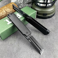 A close-up of a CRKT folding knife blade and handle, displayed next to a green box, wood log, and outdoor plants on a grey surface.