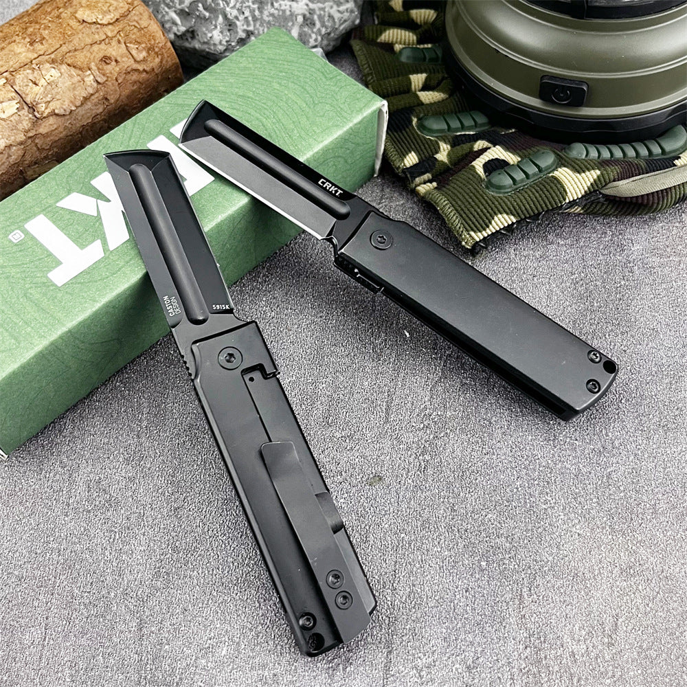 Two open black CRKT folding knives with tanto blades displayed on a grey textured surface, next to a green CRKT box, a piece of wood, green camouflage fabric, and a camping lantern.