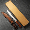 A custom fixed blade knife with a carved wooden handle and its leather sheath, displayed next to a brown kraft paper box on a dark marble-patterned surface.