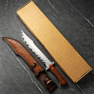 A custom fixed blade knife with a carved wooden handle and its leather sheath, displayed next to a brown kraft paper box on a dark marble-patterned surface.