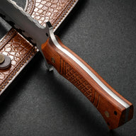 A side profile view of a custom fixed blade knife, highlighting the carved wooden handle, silver blade with black flame patterns, and the brown leather sheath.