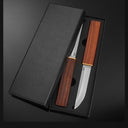 An open black gift box containing a set of D2 steel fixed blade knives with wooden handles and brass accents, displayed on a dark background.