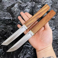 A hand holding two D2 steel fixed blade knives with wooden handles and brass accents, displayed outdoors against a rough rocky background.