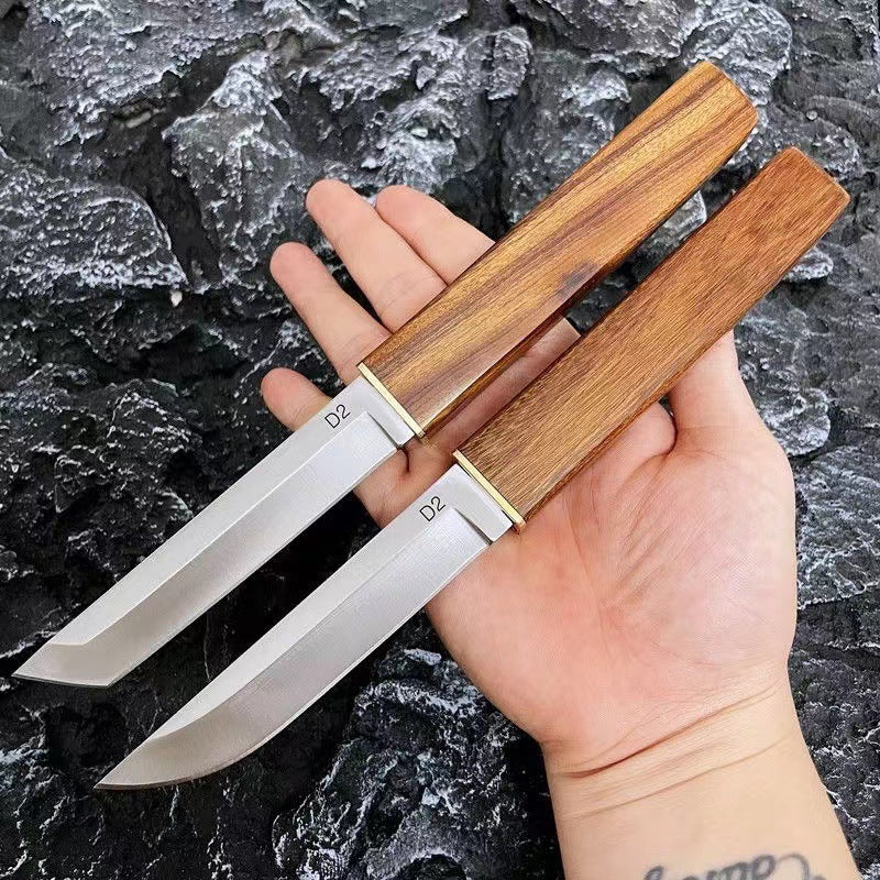 A hand holding two D2 steel fixed blade knives with wooden handles and brass accents, displayed outdoors against a rough rocky background.