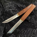 Two D2 steel fixed blade knives with wooden handles and brass accents, one with a tanto blade and one with a curved blade, displayed on a dark rocky surface.