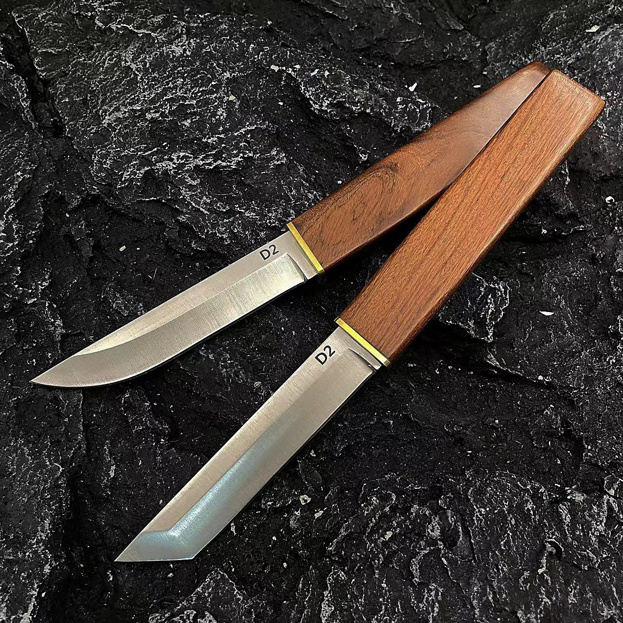 Two D2 steel fixed blade knives with wooden handles and brass accents, one with a tanto blade and one with a curved blade, displayed on a dark rocky surface.