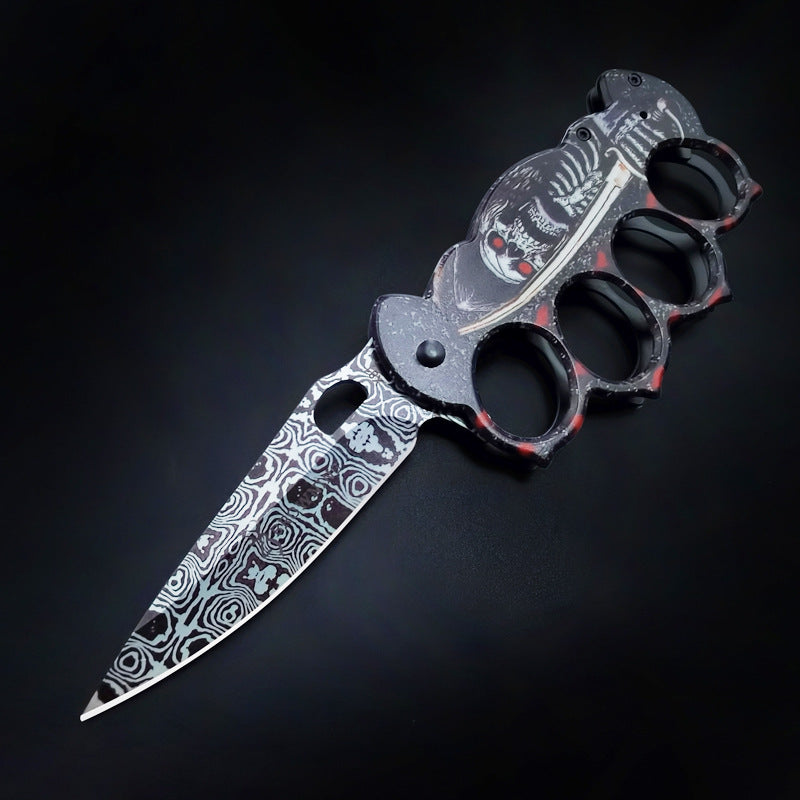 An open knuckle duster folding knife with a black and red skull graphic handle and a silver damascus-style etched blade, displayed on a dark background.