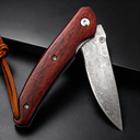 An open folding knife with a patterned Damascus steel blade and a polished red wooden handle, featuring a brown leather lanyard at the end, displayed on a dark textured background.