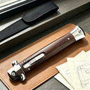 A closed stiletto knife with a dark wood handle and silver bolster, displayed on a leather notebook next to a black nylon sheath and technical sketches.