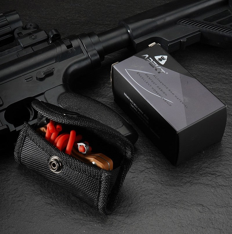 A product packaging display showing a tan EDC slingshot with red bands tucked inside a black nylon pouch, placed next to its black and silver APEXT brand packaging box, all set against a dark tactical background.