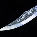 A close-up of the silver curved blade tip of a custom knife, featuring intricate tribal-style engravings, displayed on a dark background.