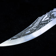 A close-up of the silver curved blade tip of a custom knife, featuring intricate tribal-style engravings, displayed on a dark background.
