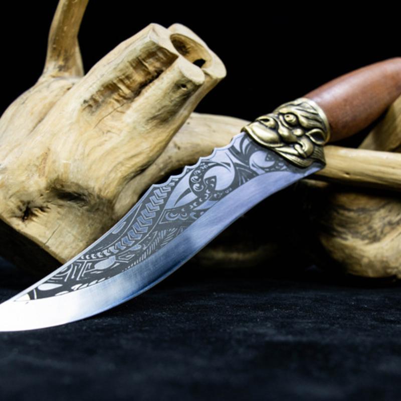 A full side view of an engraved silver curved knife with a wooden handle and brass guard, resting on a piece of driftwood against a dark background.