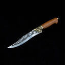A side view of an engraved curved fixed blade knife with a wooden handle, brass animal head guard, and patterned blade, displayed on a dark background.