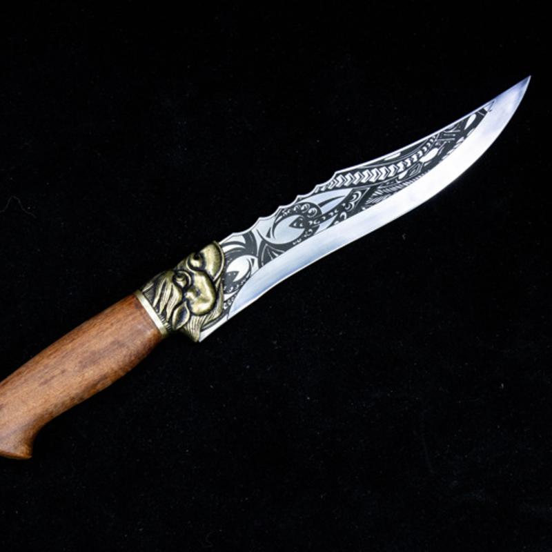 An engraved fixed blade knife featuring a curved silver blade, a wooden handle, and a brass animal head guard, displayed on a solid black background.