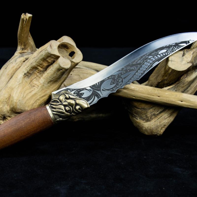 A fixed blade knife with a wooden handle, brass animal head guard, and engraved silver blade, displayed diagonally on a piece of driftwood against a dark background.