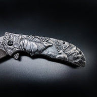 A close-up of a folding knife handle featuring detailed deep-relief engravings of a bull elk, a smaller deer, and a mountain pine landscape. The handle has black hardware and is displayed on a dark surface.