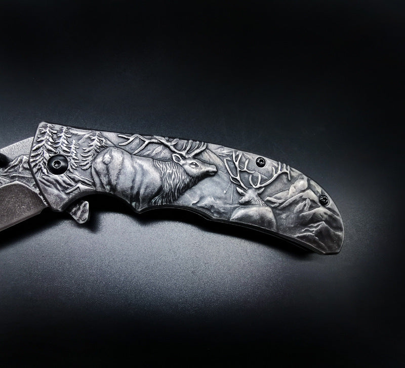 A close-up of a folding knife handle featuring detailed deep-relief engravings of a bull elk, a smaller deer, and a mountain pine landscape. The handle has black hardware and is displayed on a dark surface.
