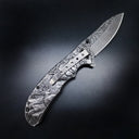 A folding knife with detailed engraved relief art of deer and a mountain forest on the handle and blade. It has a silver pocket clip and black hardware, displayed on a dark background.