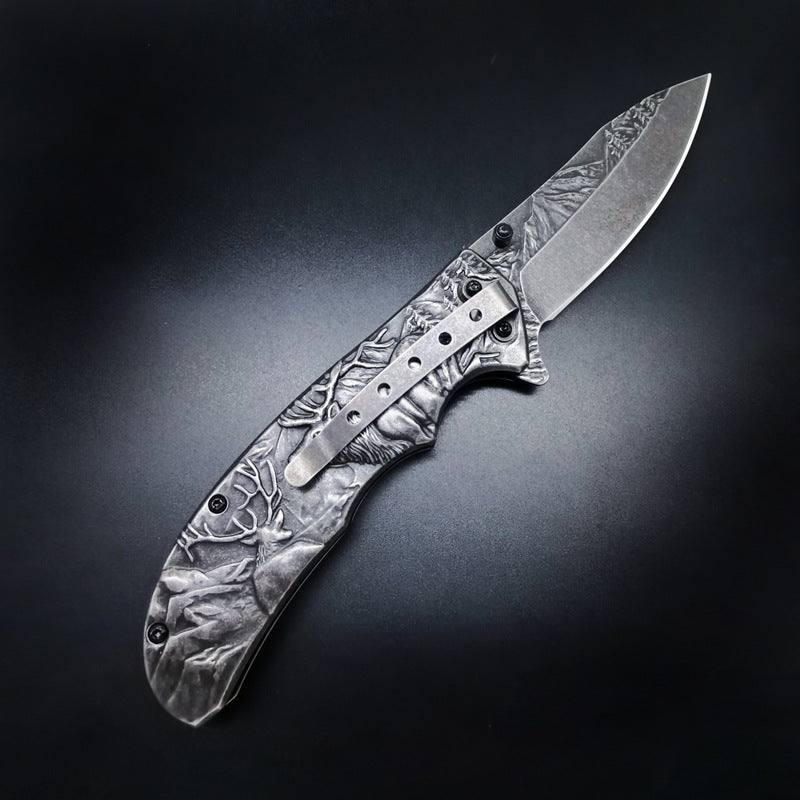 A folding knife with detailed engraved relief art of deer and a mountain forest on the handle and blade. It has a silver pocket clip and black hardware, displayed on a dark background.