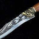 A close-up of the transition between the engraved silver blade and the brass animal head guard on a custom fixed blade knife, with a wooden handle visible.
