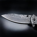 A close-up of a folding knife blade featuring a deeply engraved mountain and forest landscape on the spine, displayed on a dark background.