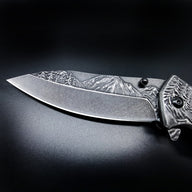A close-up of a folding knife blade featuring a deeply engraved mountain and forest landscape on the spine, displayed on a dark background.