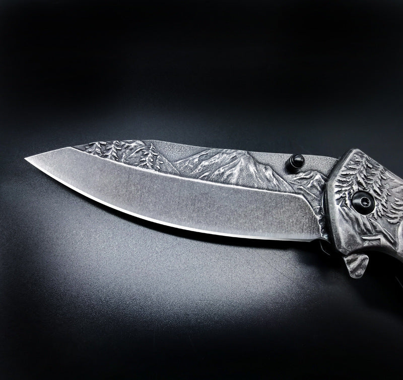 A close-up of a folding knife blade featuring a deeply engraved mountain and forest landscape on the spine, displayed on a dark background.