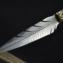 An engraved feather blade fixed knife resting on a wooden block, highlighting the detailed silver feather engraving on the blade and the hammered brass bolster, displayed on a dark background.