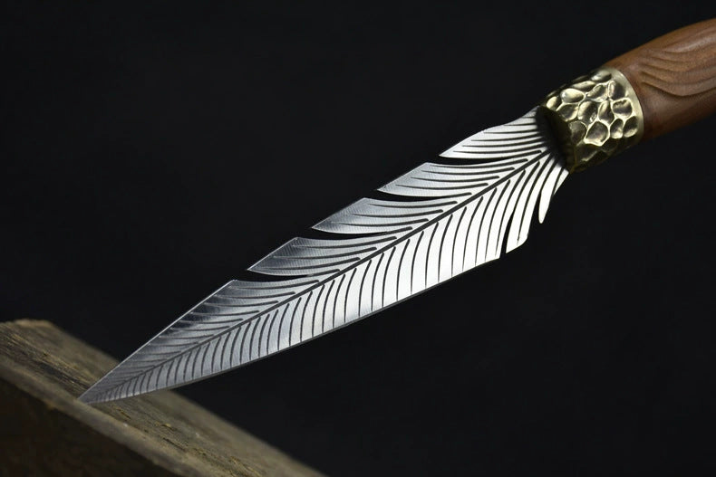 An engraved feather blade fixed knife resting on a wooden block, highlighting the detailed silver feather engraving on the blade and the hammered brass bolster, displayed on a dark background.