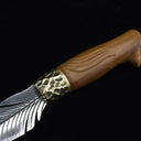 A detailed close-up of the hammered brass bolster and the carved wooden handle of an engraved feather blade fixed knife, displayed on a dark background.