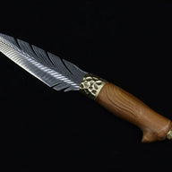 A close-up view of an engraved fixed knife featuring a silver feather blade, a hammered brass bolster, and a wooden handle, displayed on a dark background.