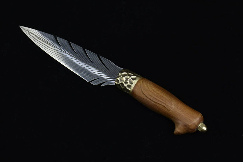 A close-up view of an engraved fixed knife featuring a silver feather blade, a hammered brass bolster, and a wooden handle, displayed on a dark background.