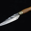 A full view of an engraved fixed knife featuring a silver feather blade, a hammered brass bolster, and a wooden handle, displayed diagonally on a dark background.