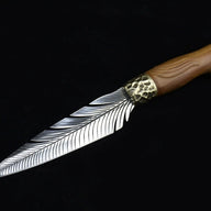 A full view of an engraved fixed knife featuring a silver feather blade, a hammered brass bolster, and a wooden handle, displayed diagonally on a dark background.