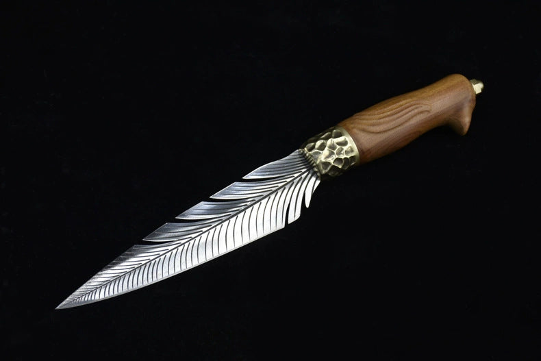 A full view of an engraved fixed knife featuring a silver feather blade, a hammered brass bolster, and a wooden handle, displayed diagonally on a dark background.