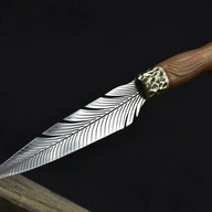 An open fixed knife with a silver feather-patterned blade and a hammered brass bolster, resting on a wooden edge against a dark background.