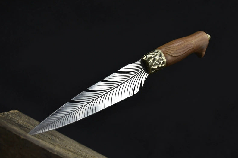 An open fixed knife with a silver feather-patterned blade and a hammered brass bolster, resting on a wooden edge against a dark background.