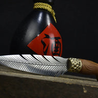 An engraved feather blade fixed knife resting horizontally in front of a black liquor gourd with a red label, displayed on a dark wooden surface.