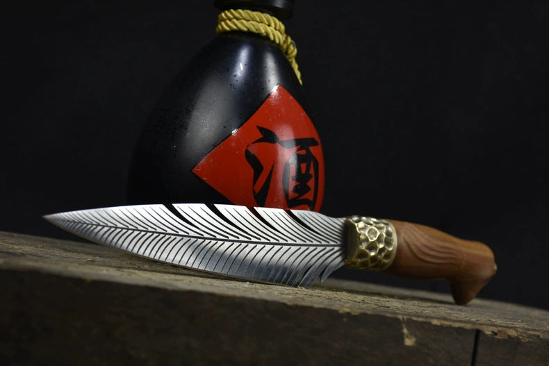 An engraved feather blade fixed knife resting horizontally in front of a black liquor gourd with a red label, displayed on a dark wooden surface.