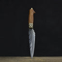 An engraved feather blade fixed knife standing vertically on a wooden surface, showcasing the wooden handle, hammered brass bolster, and silver blade against a dark background.