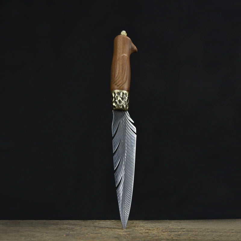 An engraved feather blade fixed knife standing vertically on a wooden surface, showcasing the wooden handle, hammered brass bolster, and silver blade against a dark background.