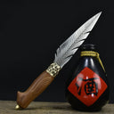 An engraved feather blade fixed knife with a wooden handle and brass bolster, displayed diagonally next to a black liquor gourd with a red diamond label on a dark background.