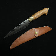 An engraved feather blade fixed knife with a wooden handle and a hammered brass bolster, displayed diagonally next to a brown leather sheath on a dark background.