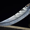 A close-up of the curved silver blade of a custom fixed knife, highlighting the intricate feather engraving, displayed next to a black liquor gourd.