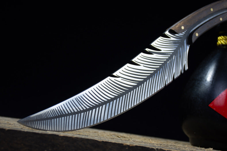 A close-up of the curved silver blade of a custom fixed knife, highlighting the intricate feather engraving, displayed next to a black liquor gourd.